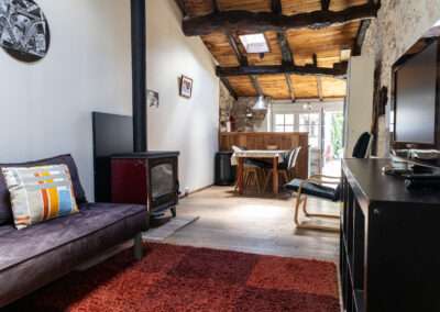 Cozy rustic living space with a wood-burning stove, exposed ceiling beams, and a purple sofa with a colorful striped pillow, leading to a dining area.