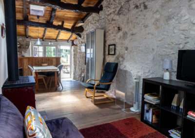 Rustic living space with stone walls, wooden beams, a wood stove on the left, and a dining area beyond a door in back.