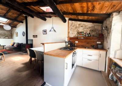 Rustic attic-style kitchen and living area with exposed dark wooden beams, stone wall, and white cabinetry; island with a gas stove and a seating area nearby.